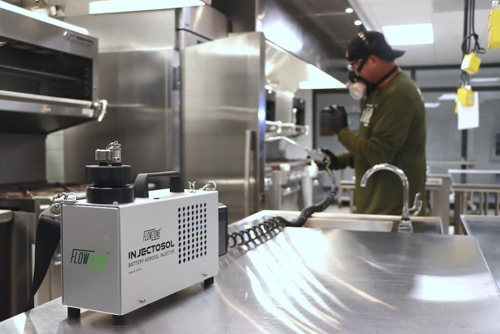 Person in a kitchen wearing a gas mask with a device labeled 'FlowZone Injectosol' on a stainless steel counter.
